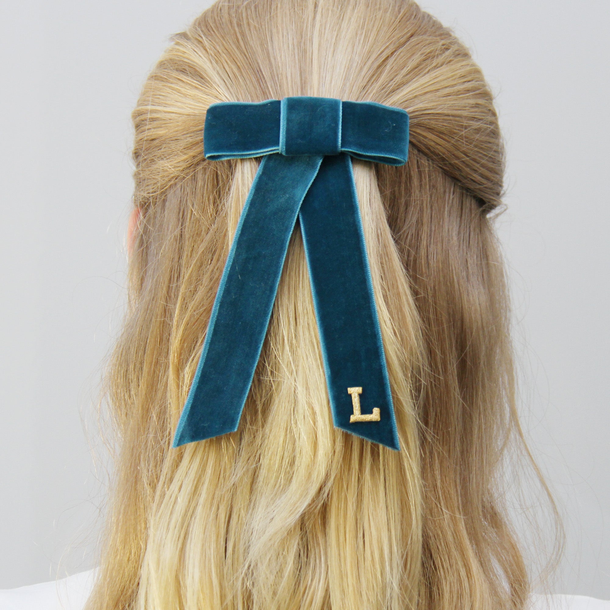 Monogrammed teal velvet hair bow with a gold letter 'L' on a person with blonde hair.