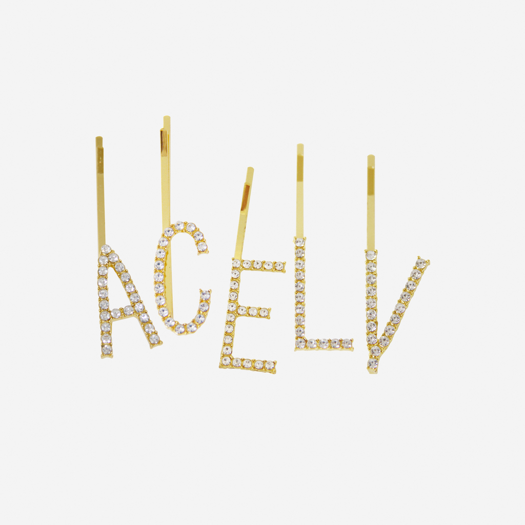 Five crystal and gold hair slides with the initials 'A' C' 'E' 'L' and 'V' on an off white background