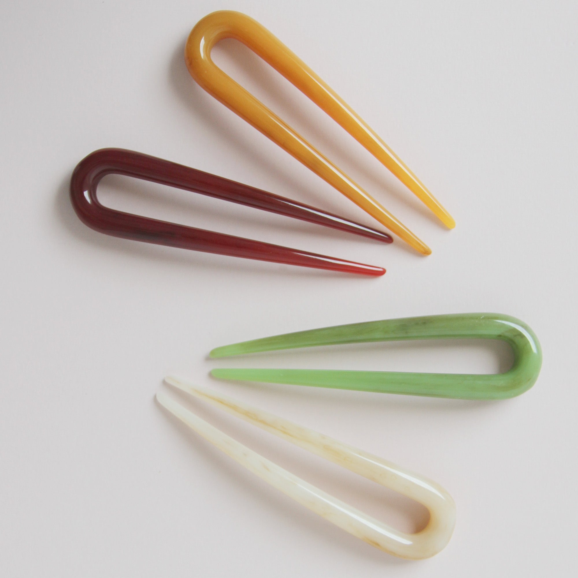 Set of four colorful resin hair pins on a light pink background, hair pins from top to bottom are amber, burgundy, green, cream