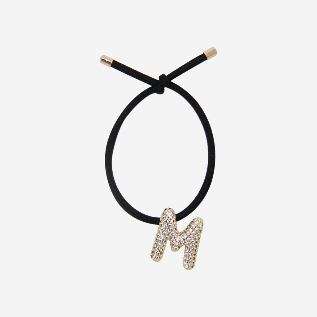 Black hair tie with gold and crystal letter 'M' charm on an off white background