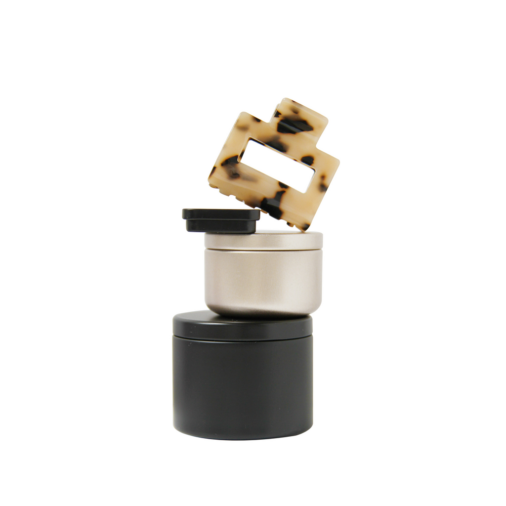 Braid & Bow gift set, stack of black and gold tins containing hair claws, hair ties and kirby grips with a tortoiseshell hair claw balanced on top, on a white background