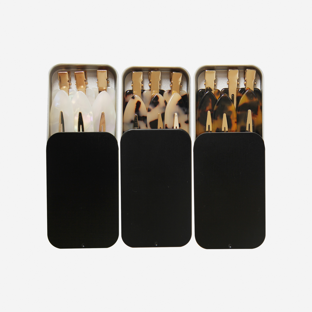 Three black tins containing sets of 6 non crease hair clips in the colours mother of pearl, light tortoiseshell and dark tortoiseshell on an off white background
