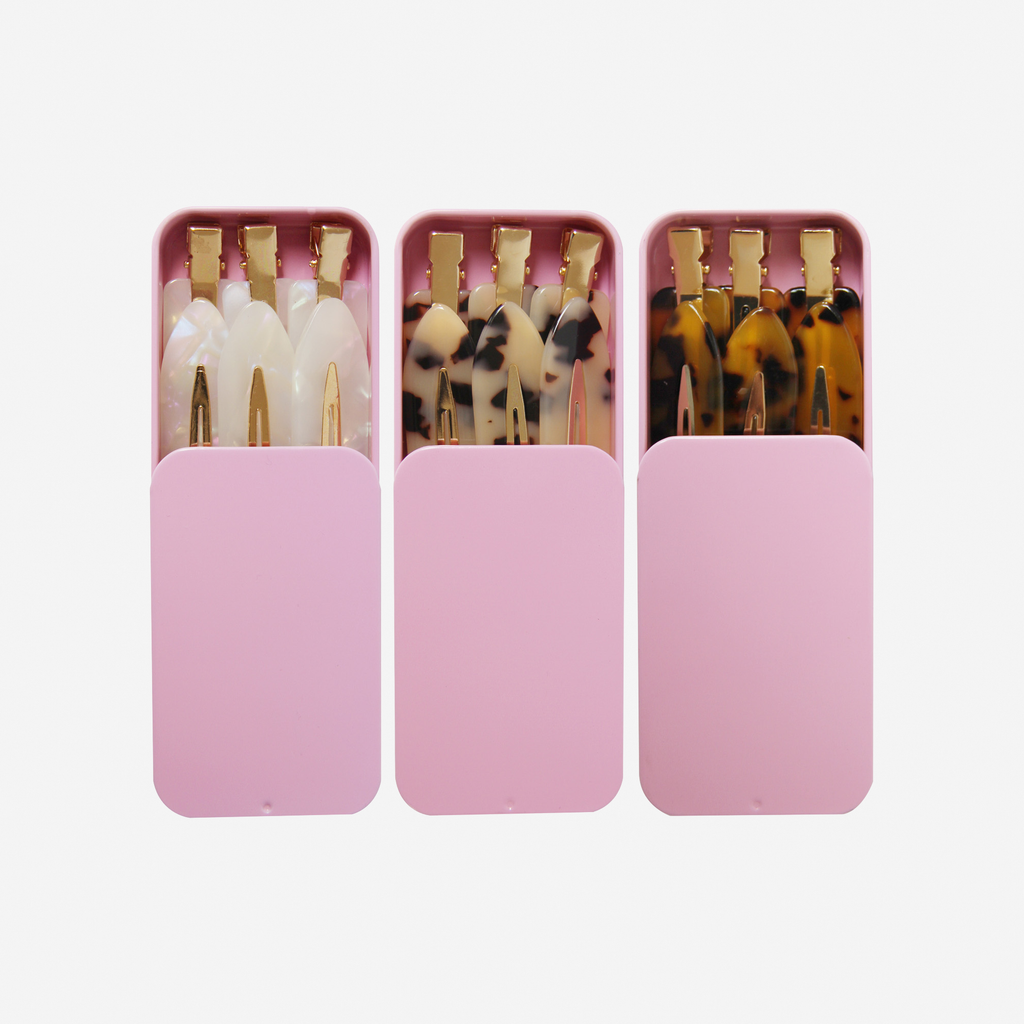 Three pink tins with non crease hair clips in them of three different colours, on an off white background