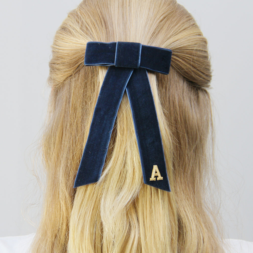 Monogrammed navy blue velvet hair bow with a gold letter 'A' on blonde hair against a light grey background