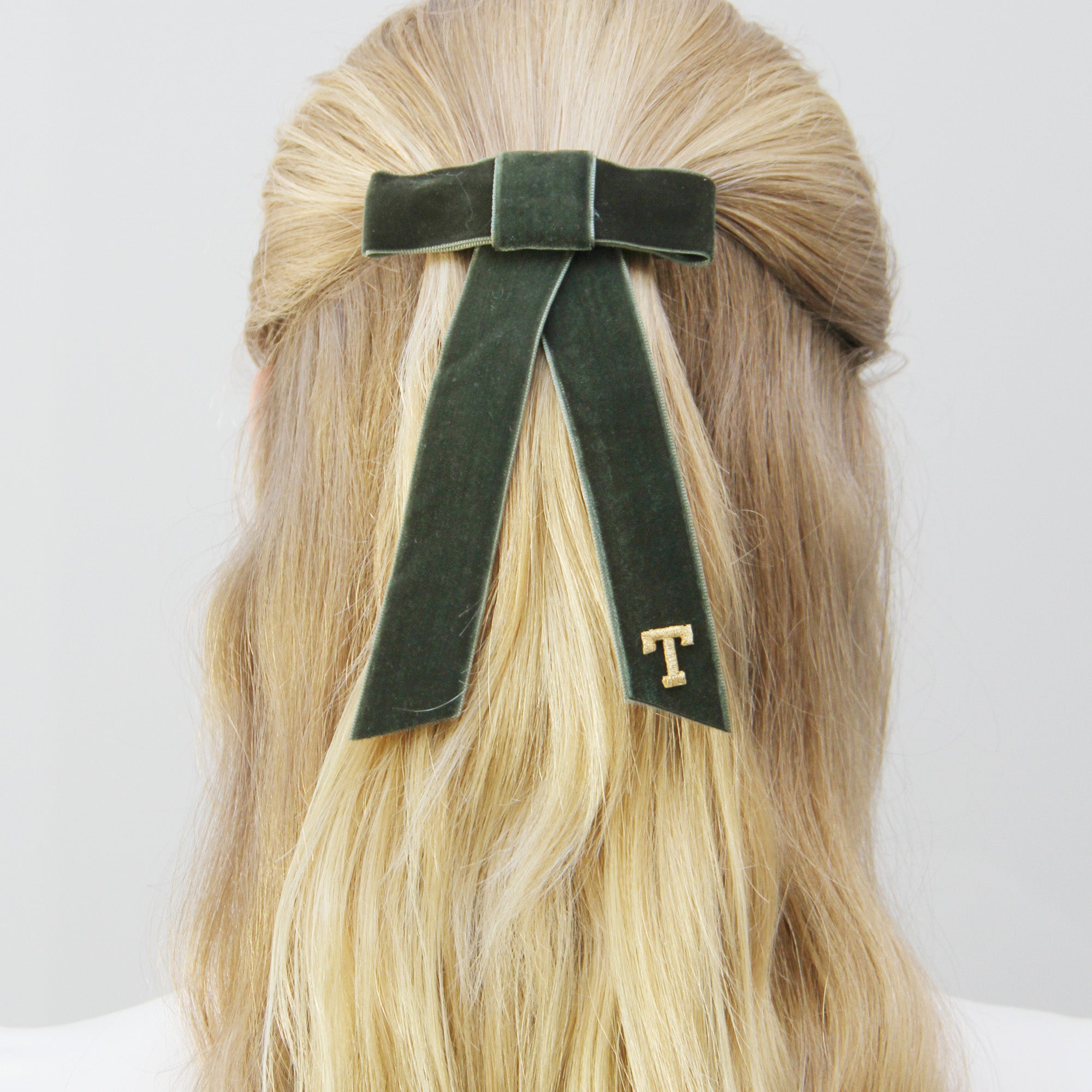Monogrammed green velvet hair bow with a gold 'T' on blonde hair against a white background