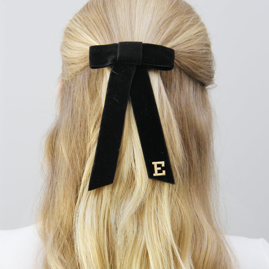 Monogrammed black hair bow with a gold letter 'E' on blonde hair against a white background