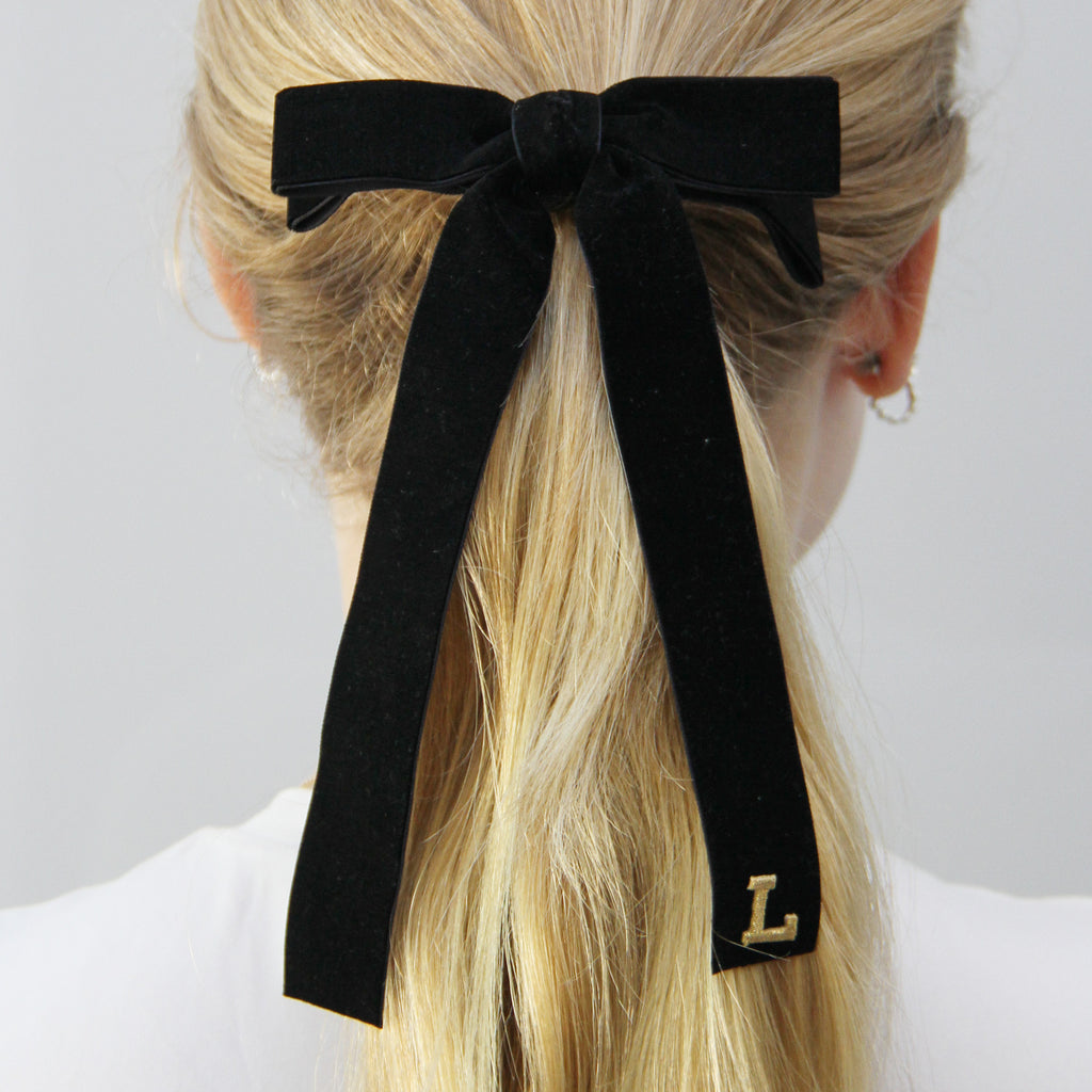 Black velvet bow hair tie with a gold letter 'L' being worn in a ponytail on a model with long blonde hair