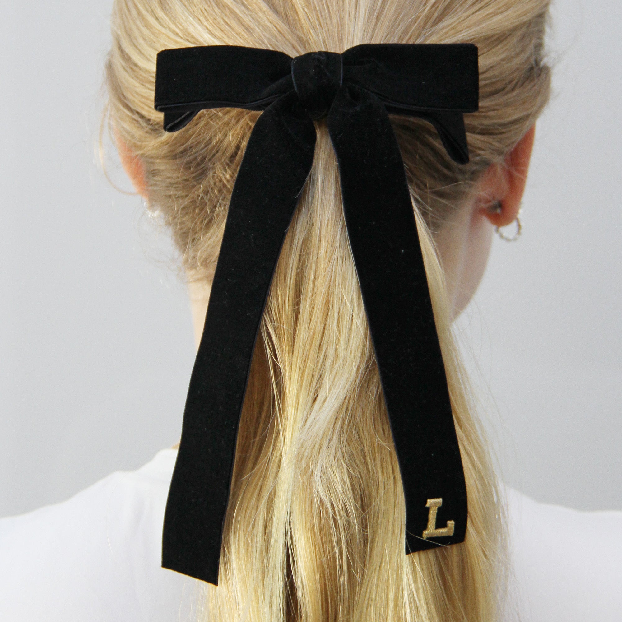 Black velvet bow hair tie with a gold letter 'L' being worn in a ponytail on a model with long blonde hair