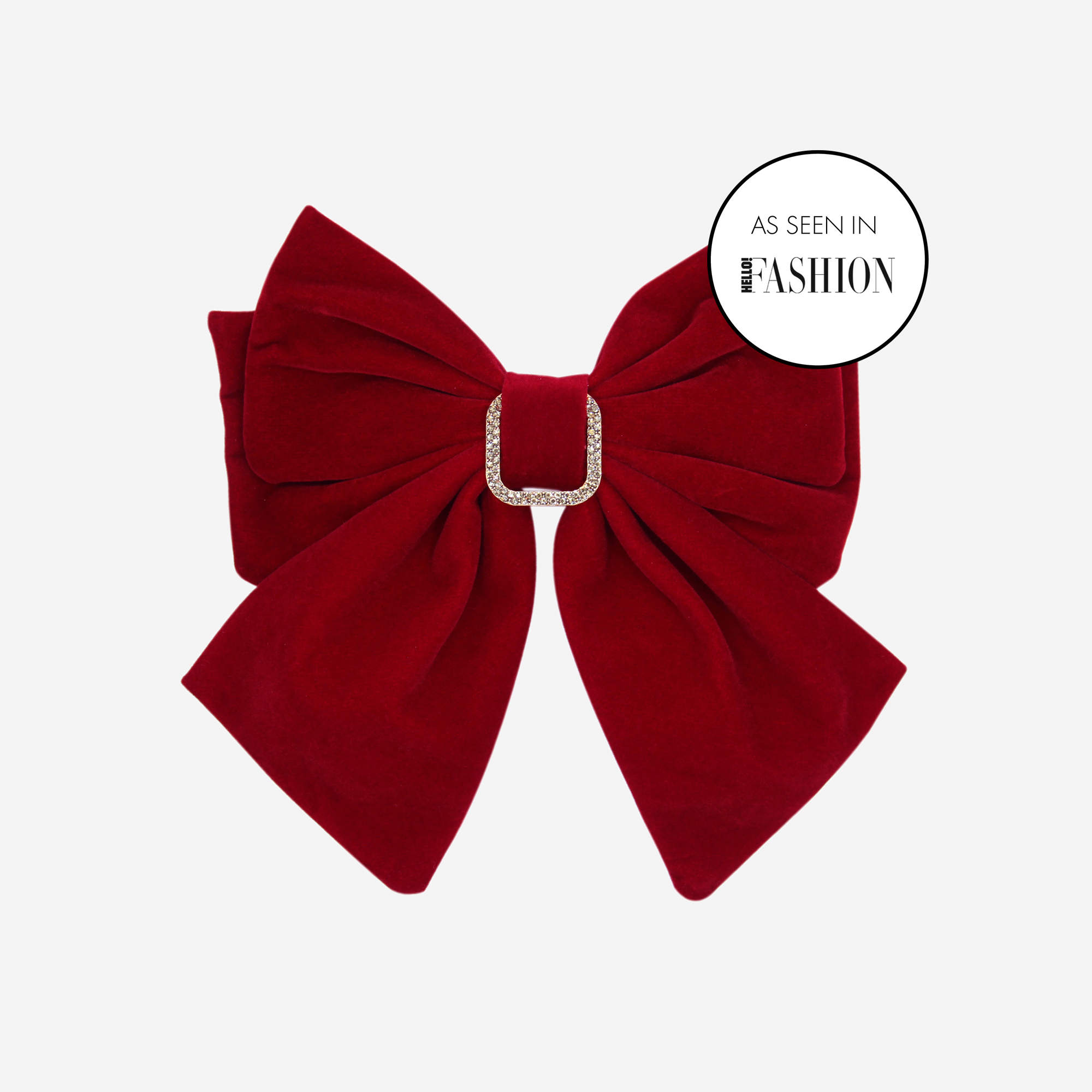 Red velvet hair bow with crystal detail in the center on an off white background and a label sating as seen in Hello Fashion