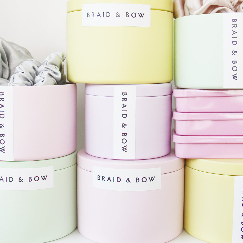 Pastel-colored tins with 'Braid & Bow' branding on a white background